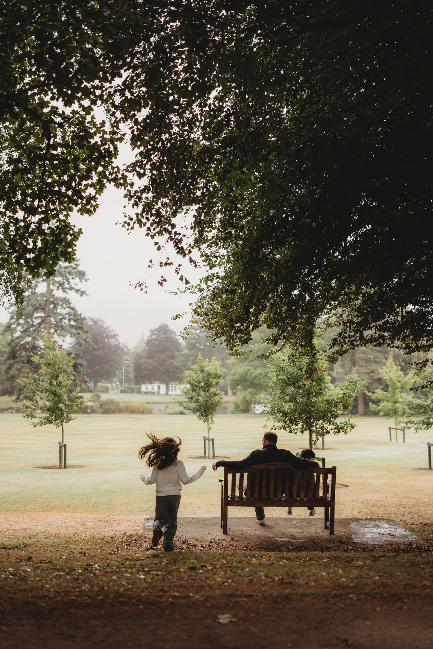 A peaceful scene in a park with figures sitting on a bench while a child plays under autumn trees.