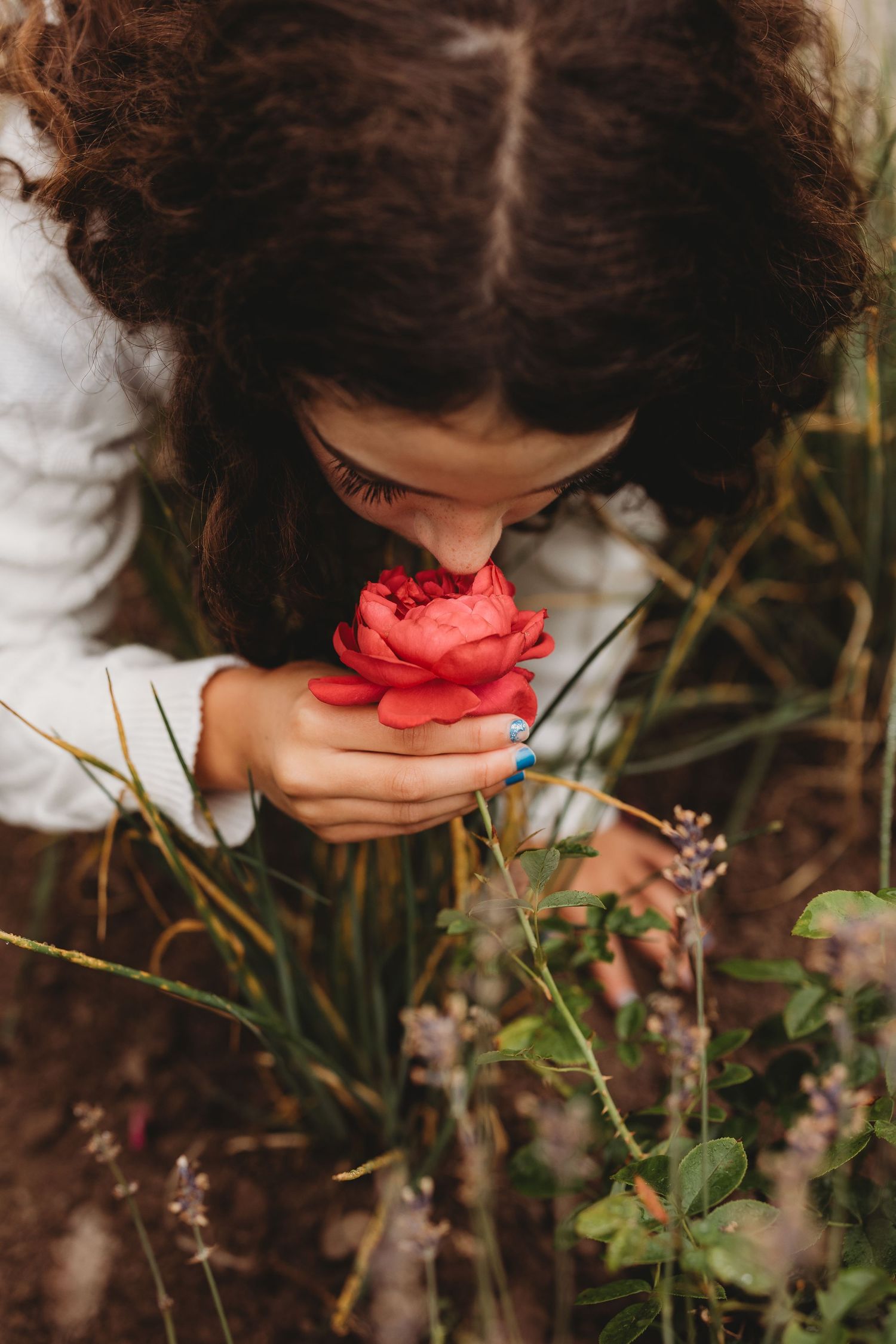 A person with dark curly hair bends down to smell a vibrant red rose in a garden setting.