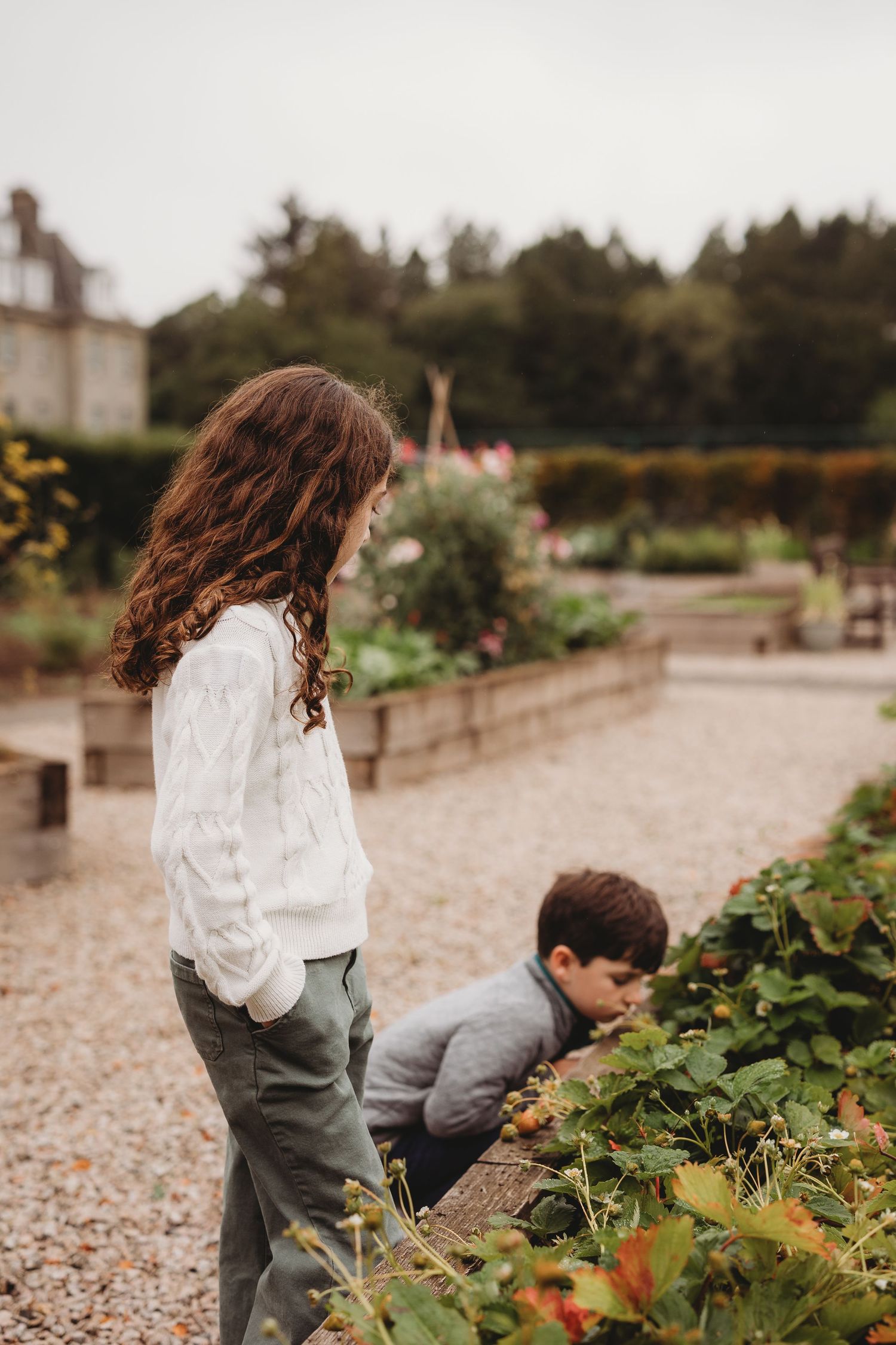 Children exploring and playing in garden area with plants and gravel path on cloudy day.