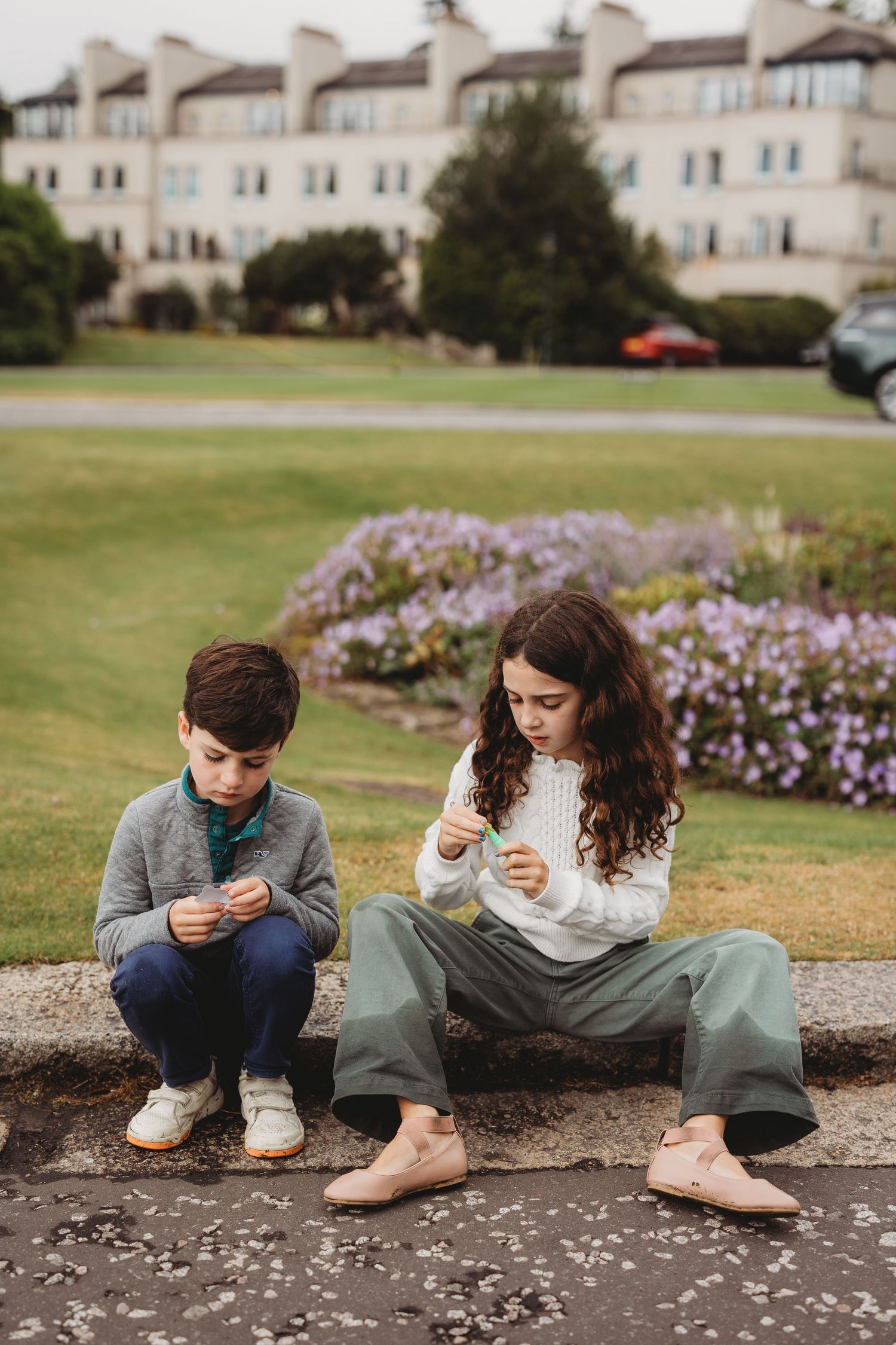 Two people sit on a stone ledge in front of a grassy lawn and building while looking at their phones.
