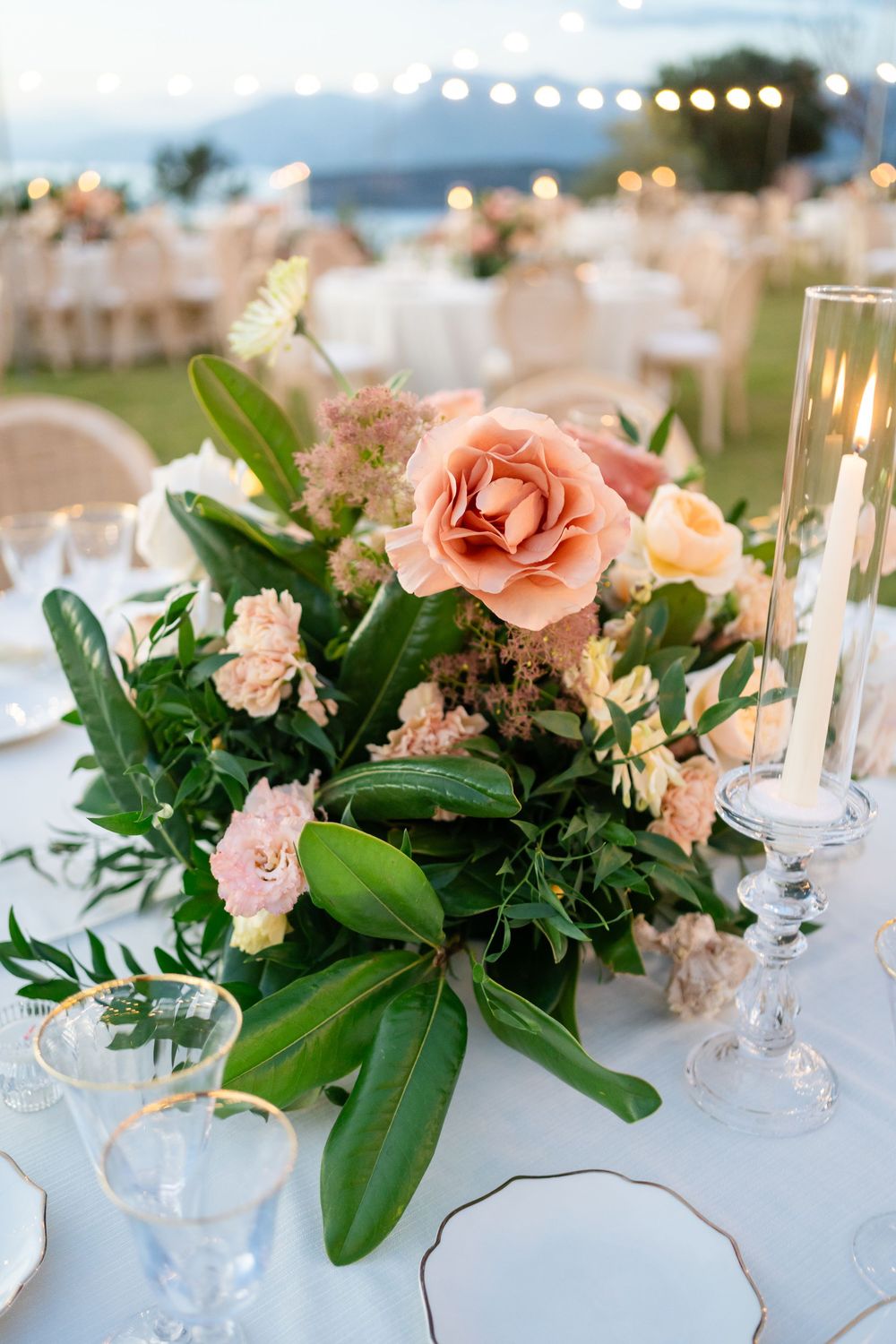 Elegant wedding centerpiece with peach roses and lush greenery on white tablecloth with glowing candle at outdoor reception.