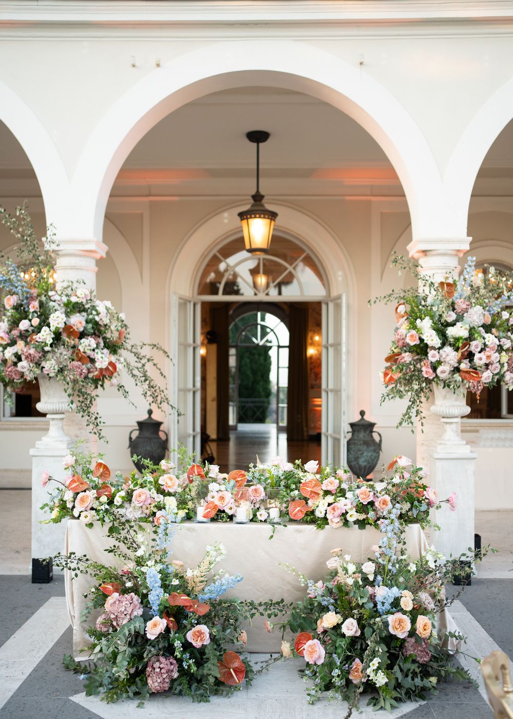 Elegant floral arrangements with pink and white blooms adorn a series of arched columns in a classic architectural setting.