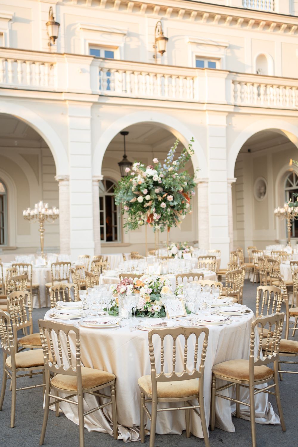 Elegant wedding reception venue with white arches and columns featuring round tables with gold chairs and floral centerpieces.