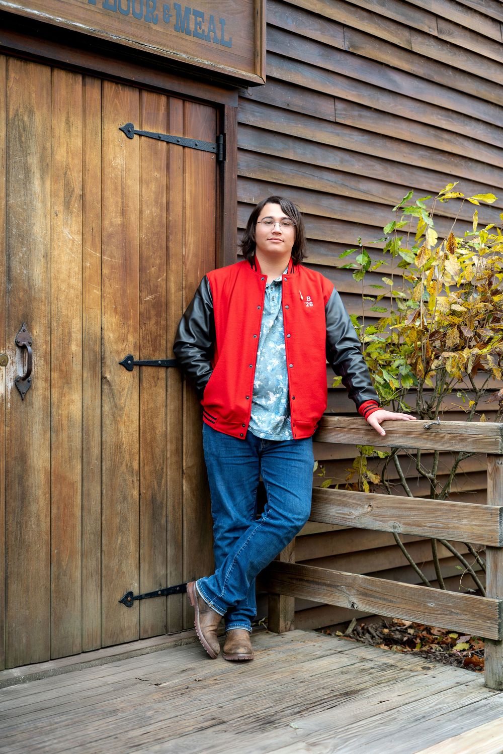 Young person in red varsity jacket and jeans leaning against rustic wooden door outdoors.