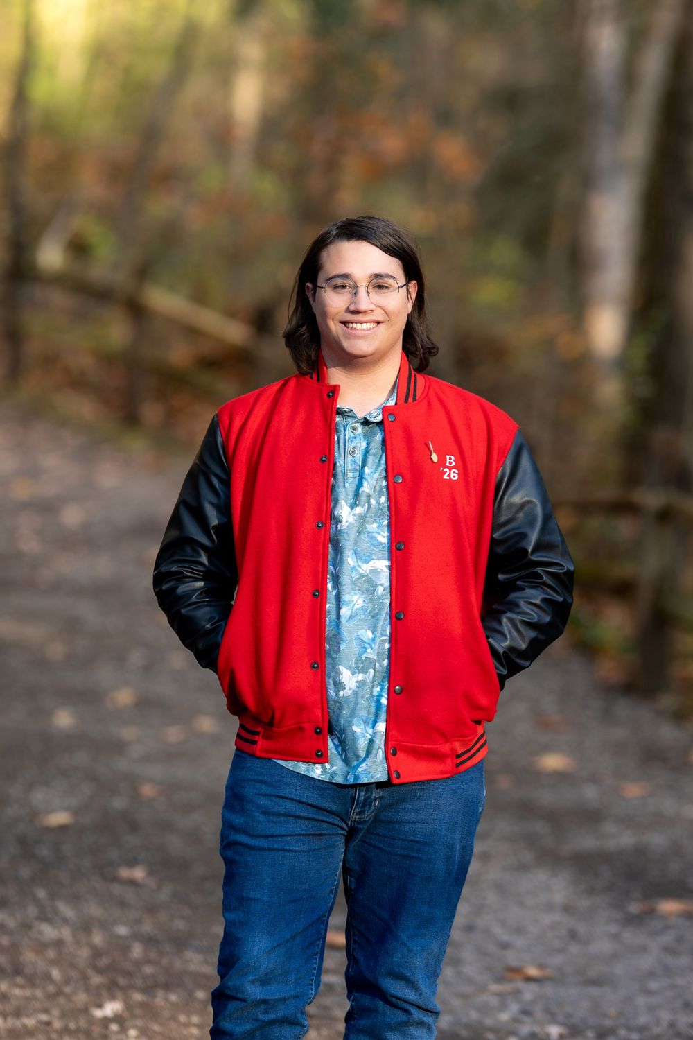 Smiling teen in red varsity jacket and blue floral shirt standing on autumn forest path.