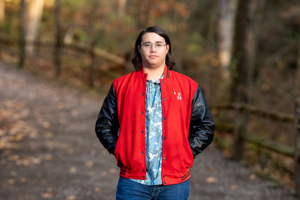 Teen in red and black varsity jacket posing seriously on a wooded trail in fall.
