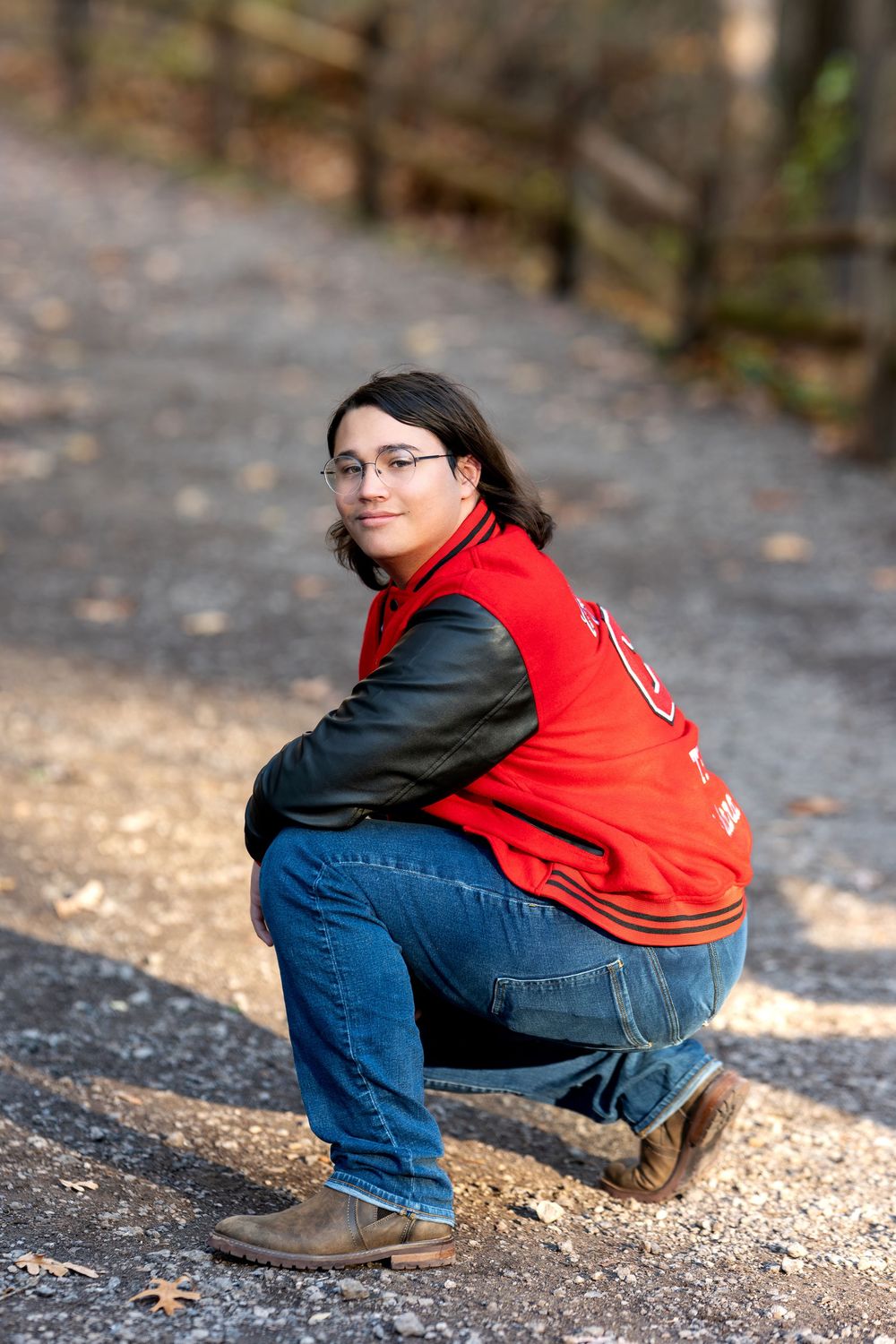 Teen in red varsity jacket crouching and smiling on a leaf-covered woodland path.