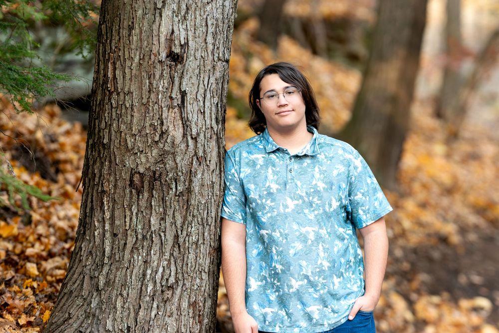 Teen in blue floral shirt leaning against a tree trunk surrounded by fall foliage.