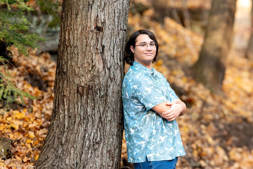 Teen in blue floral shirt leaning against tree with arms crossed in fall forest.