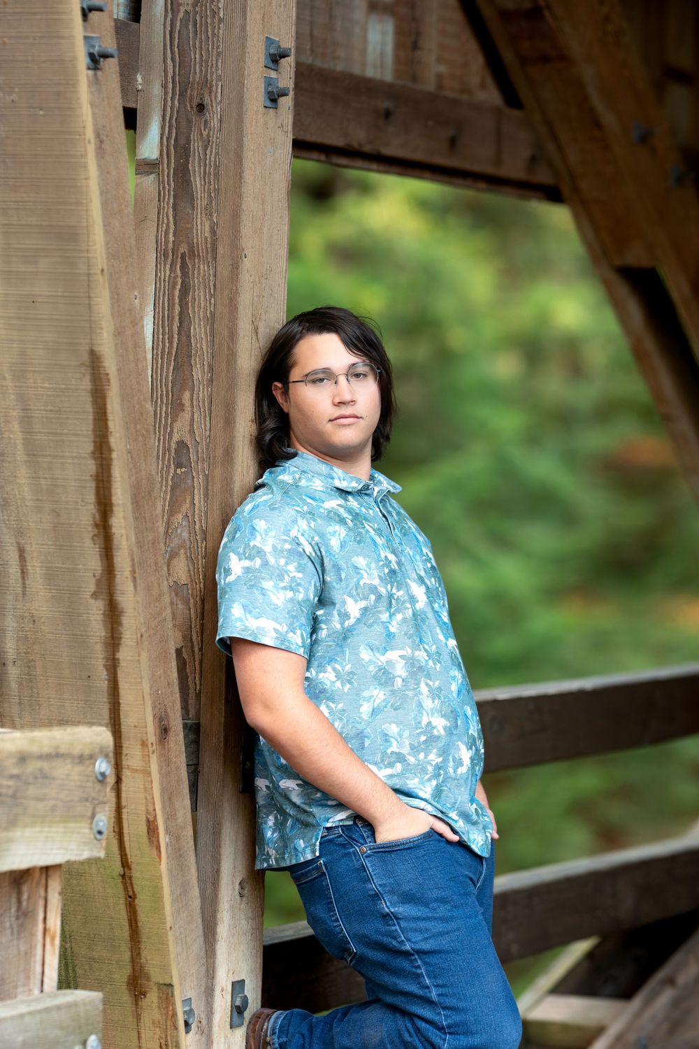 Teen boy in blue floral shirt leaning against wooden covered bridge post with hands in pockets.
