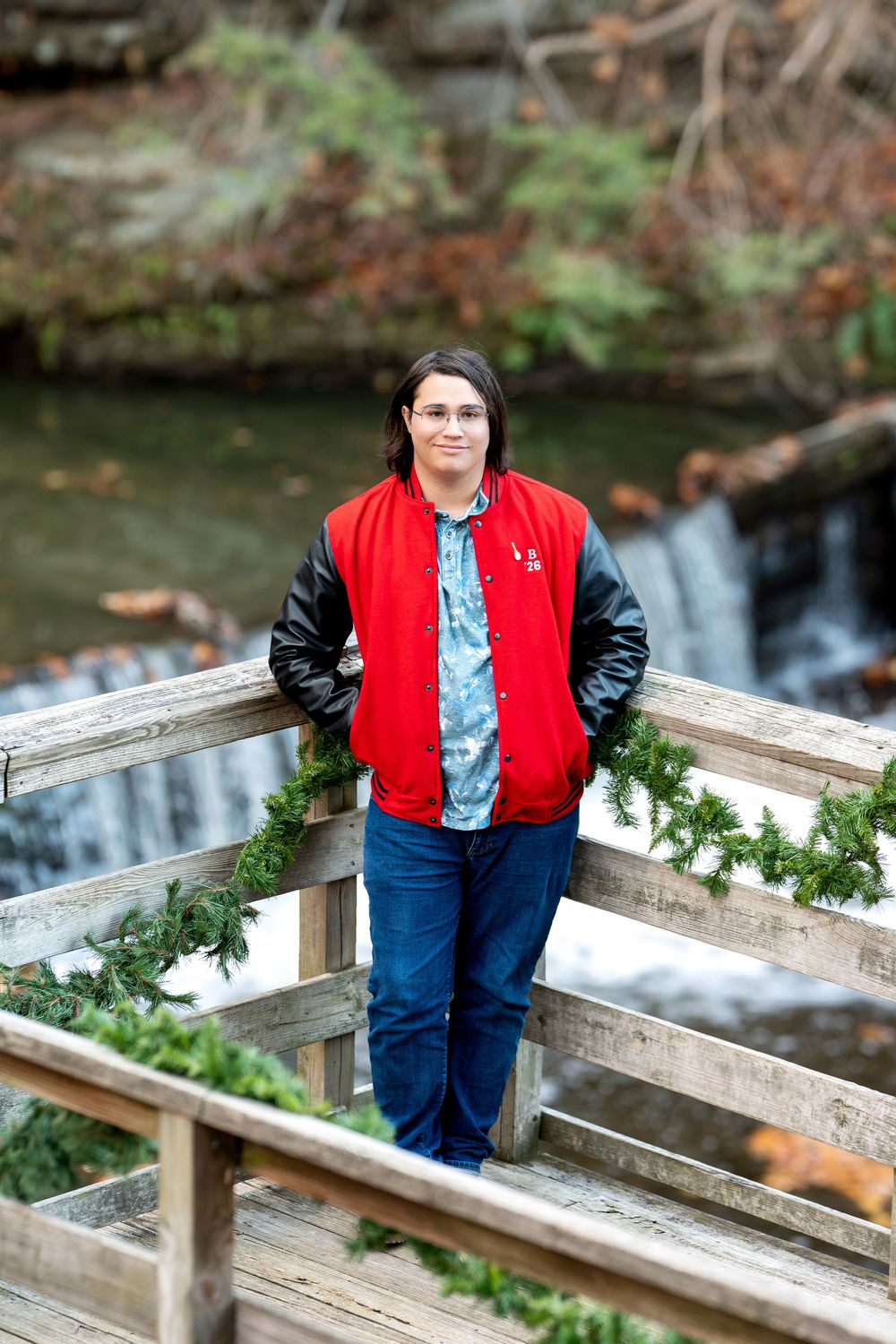 Teen boy in red varsity jacket standing on wooden bridge with holiday garland and waterfall.