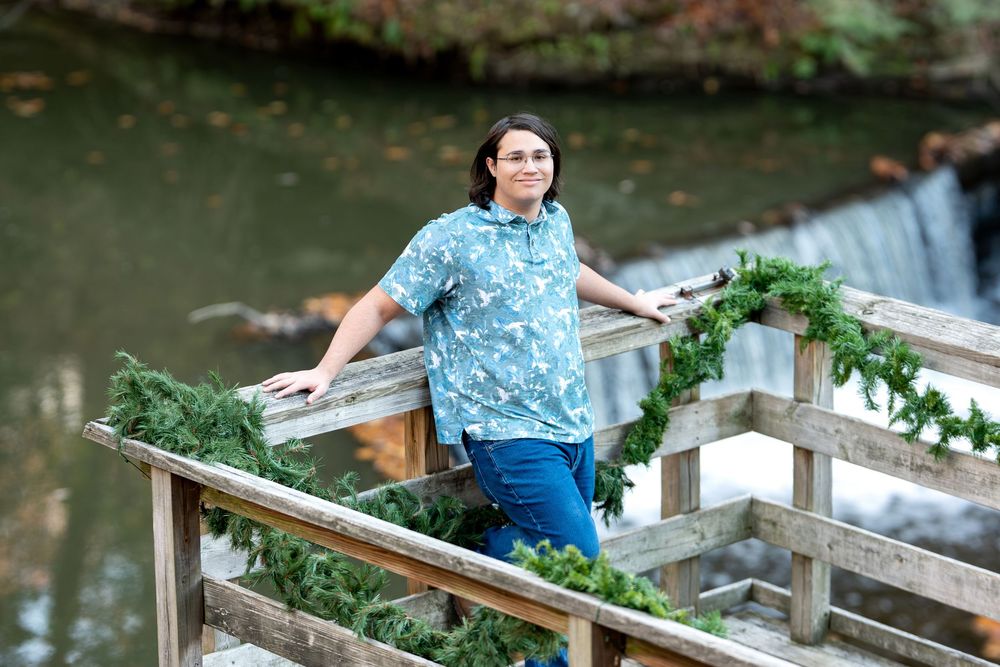 Young man in blue floral shirt smiling on wooden bridge with waterfall in background.