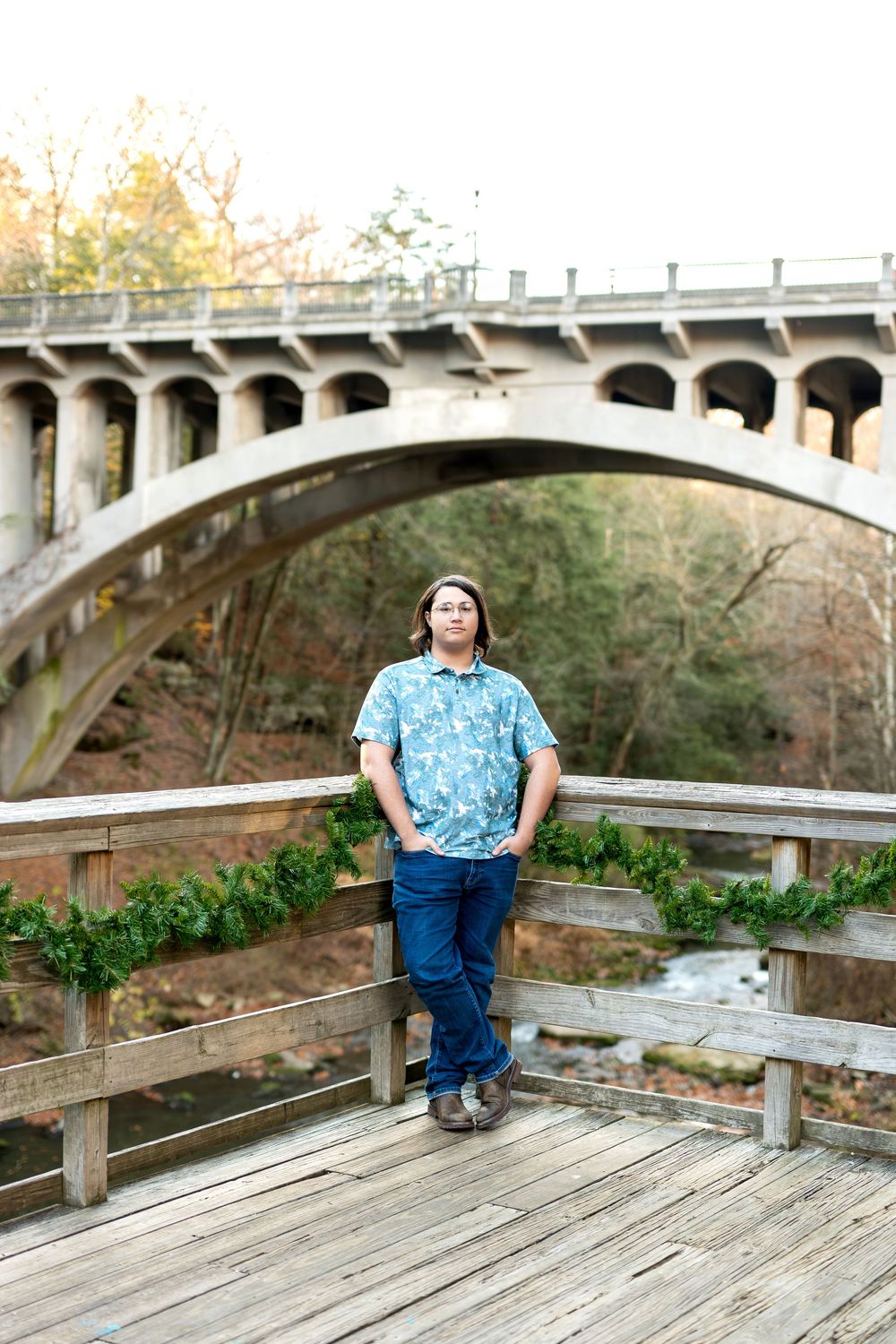 Teen boy in blue shirt standing on wooden deck with large stone arch bridge behind him.
