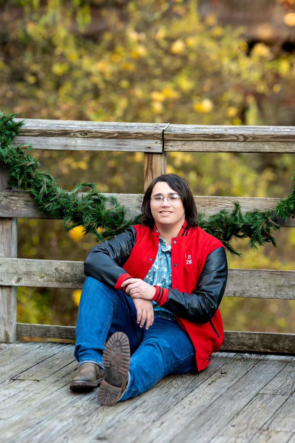 Teen in red varsity jacket sitting on wooden bridge floor with green garland backdrop.