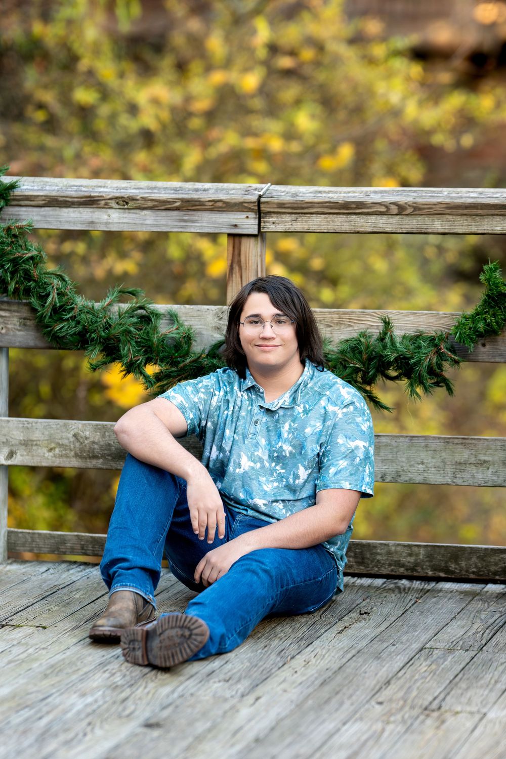 Young man in blue tie-dye shirt sitting relaxed on wooden bridge deck with fall foliage.
