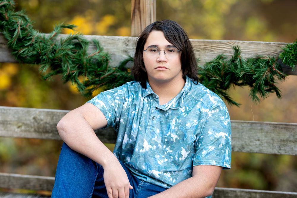Teen boy in blue splatter-paint shirt sitting against wooden fence with green garland.