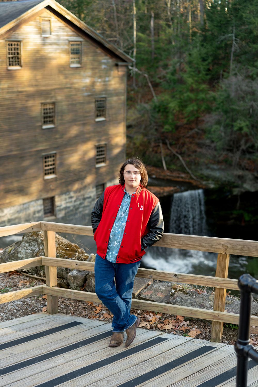 Teen in red varsity jacket stands on deck near historic mill and waterfall.