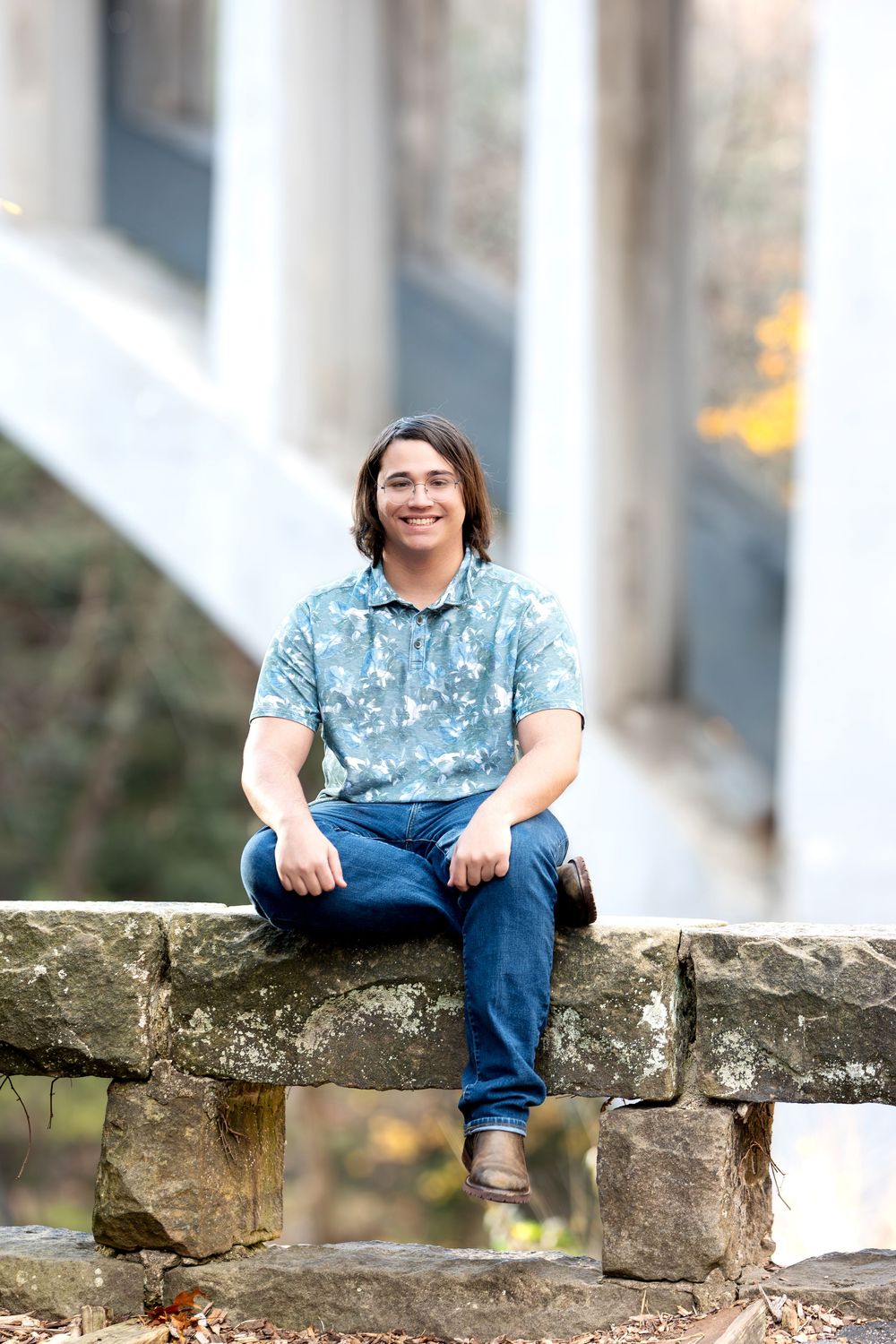 Smiling teen in floral blue polo shirt sitting cross-legged on stone wall outdoors.