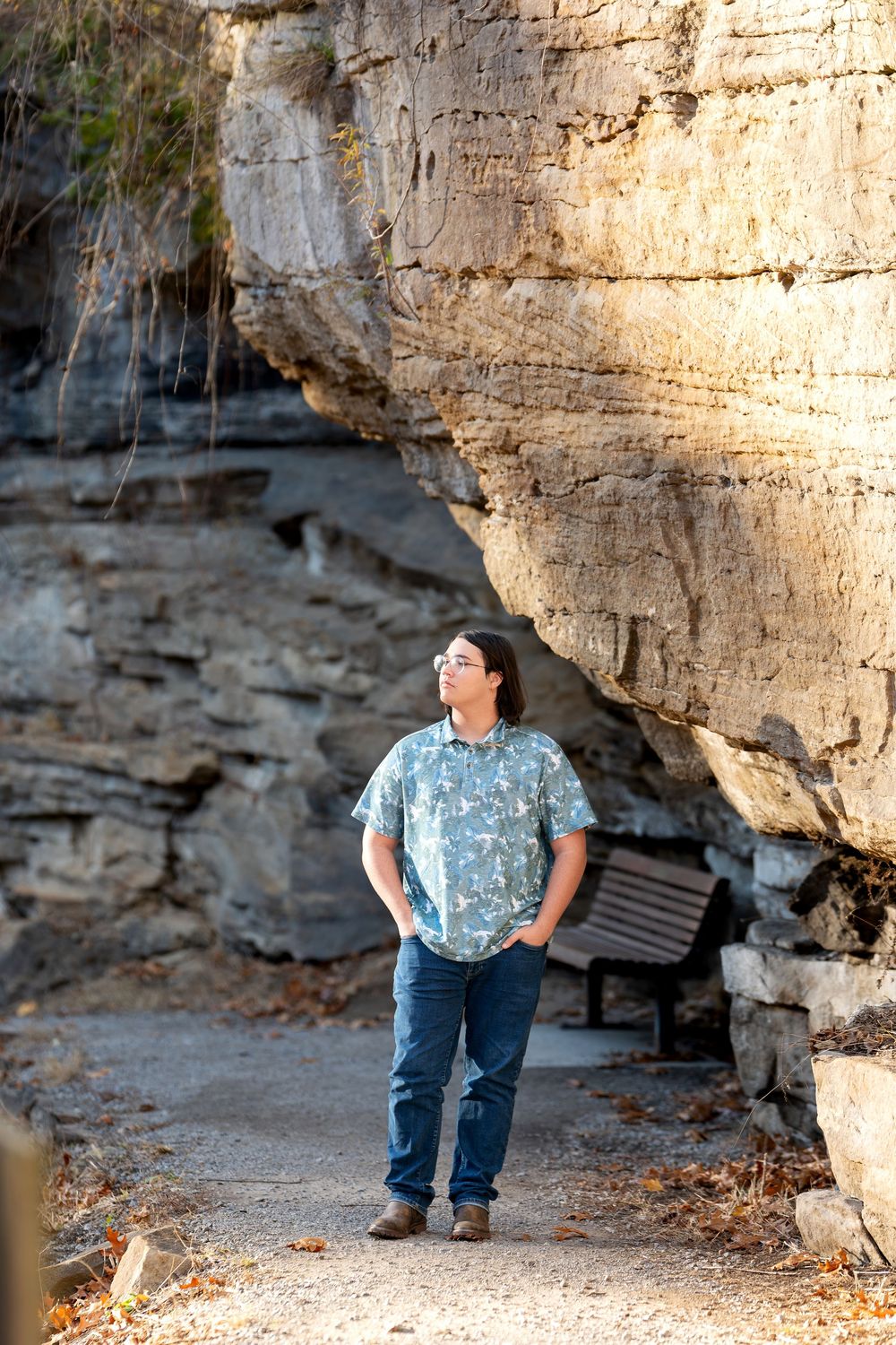 Young person in floral shirt standing on path before large natural rock cliff formation.