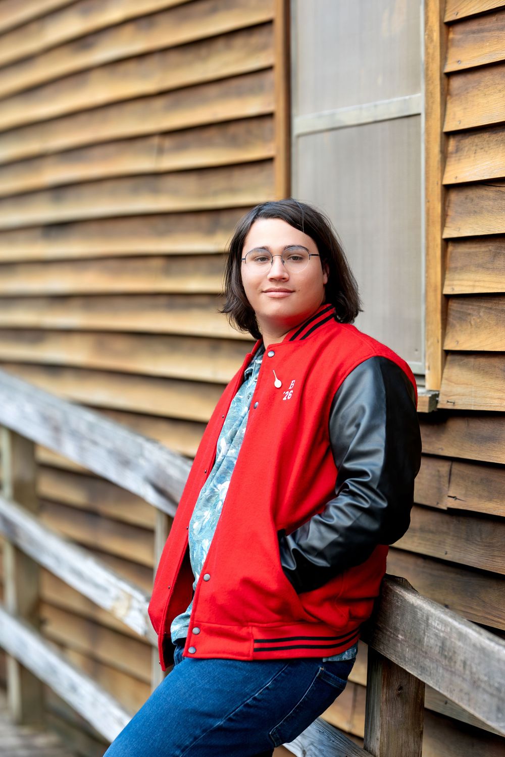 Smiling teen in red and black varsity jacket leaning against wooden slatted wall outdoors.