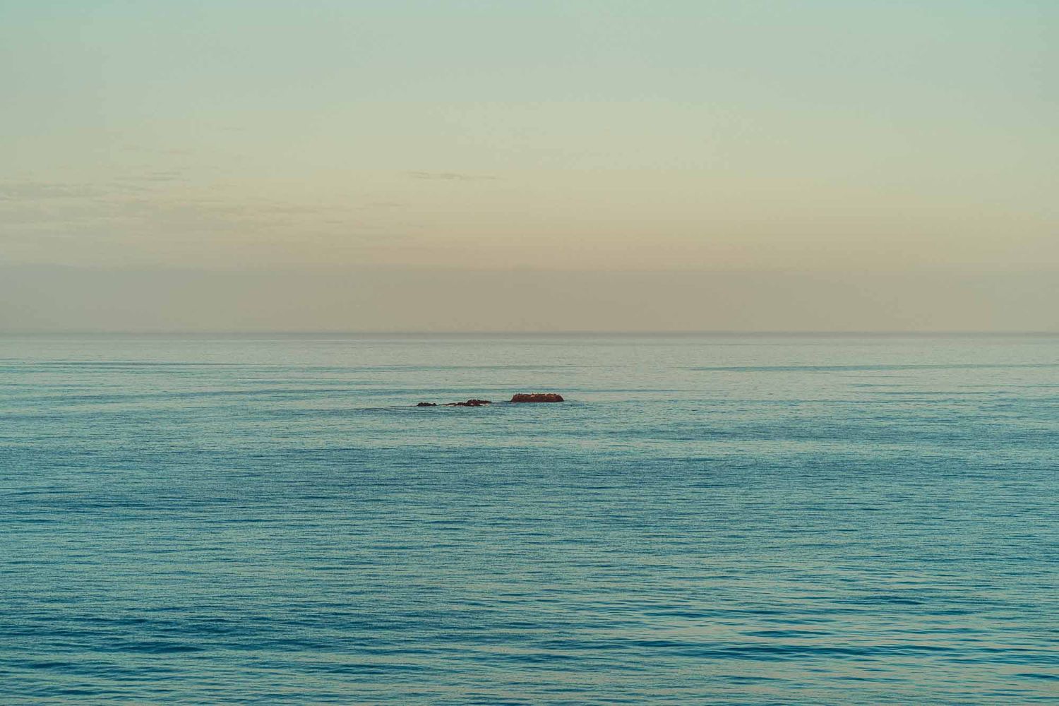 Tranquil ocean landscape with small rock formations visible on horizon during sunset against turquoise waters.