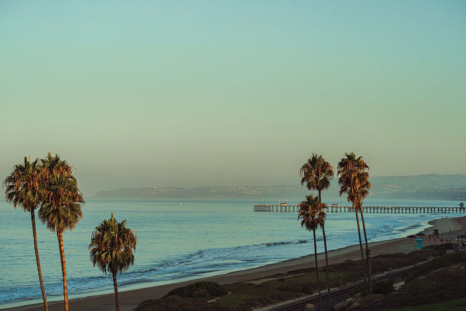 Scenic coastal view of palm trees and a long pier stretching into the ocean at sunset in Southern California.