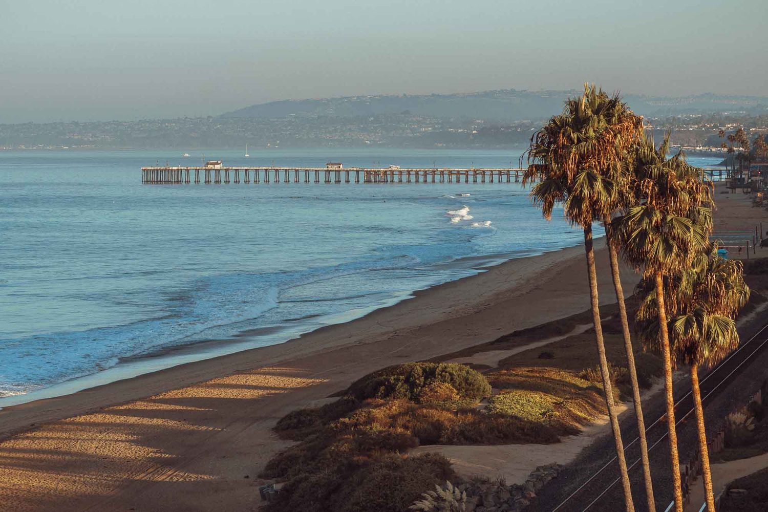 A scenic coastal view with tall palm trees overlooking a beach pier and mountains in the distance during sunset.