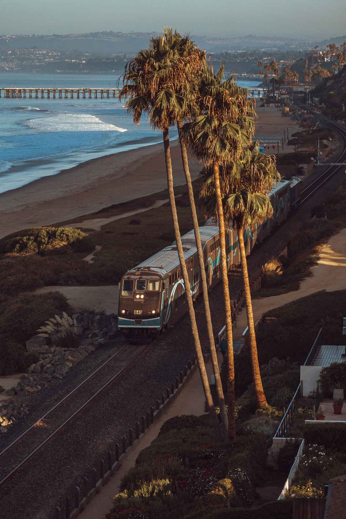 Coastal landscape with tall palm trees overlooking a beach and pier at sunset.
