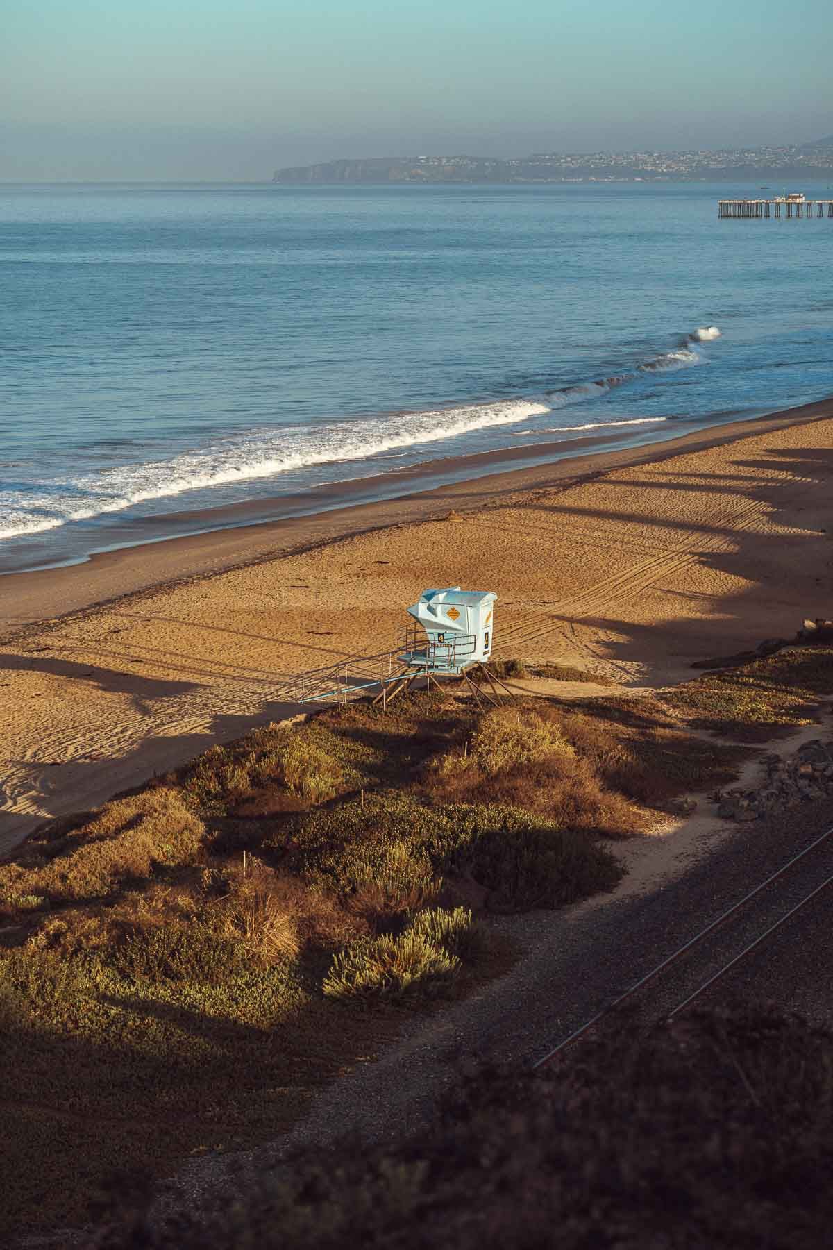 Aerial panoramic view of a sandy beach with ocean waves and distant cliffs during golden hour.