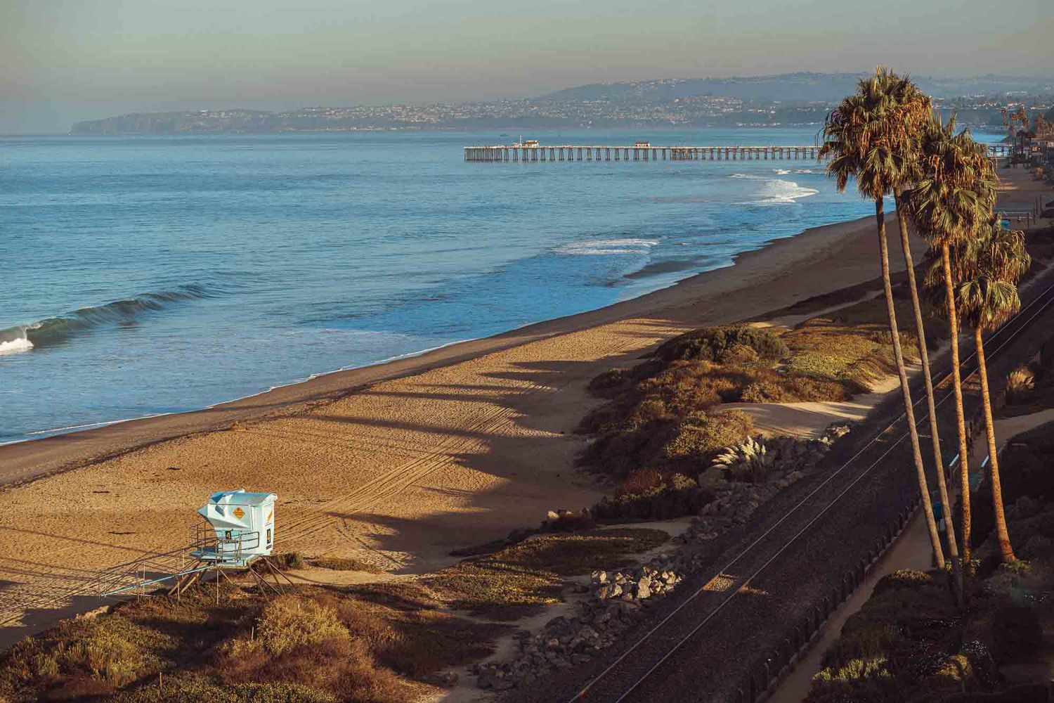 Coastal landscape with palm trees and lifeguard tower overlooking a wide sandy beach at sunset.