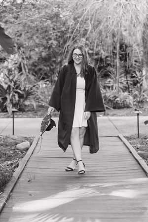 A person in a black blazer and white dress walks along a wooden boardwalk surrounded by lush greenery.