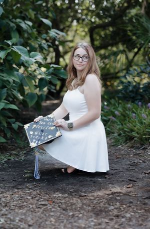 A white dress-clad figure sits on the ground holding a decorative sign in a garden setting.