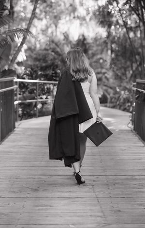 A figure in graduation attire walking away on a wooden boardwalk in black and white.