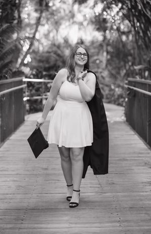 Graduate in white dress and black gown poses on wooden boardwalk surrounded by lush greenery.