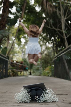 Graduate jumps joyfully on wooden bridge with graduation cap and flowers in foreground.