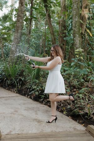 Graduate in white dress sprays champagne while celebrating on wooden pathway.