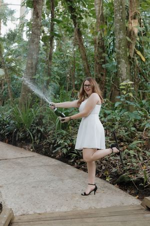 Graduate sprays champagne while celebrating on wooden pathway surrounded by tropical plants.