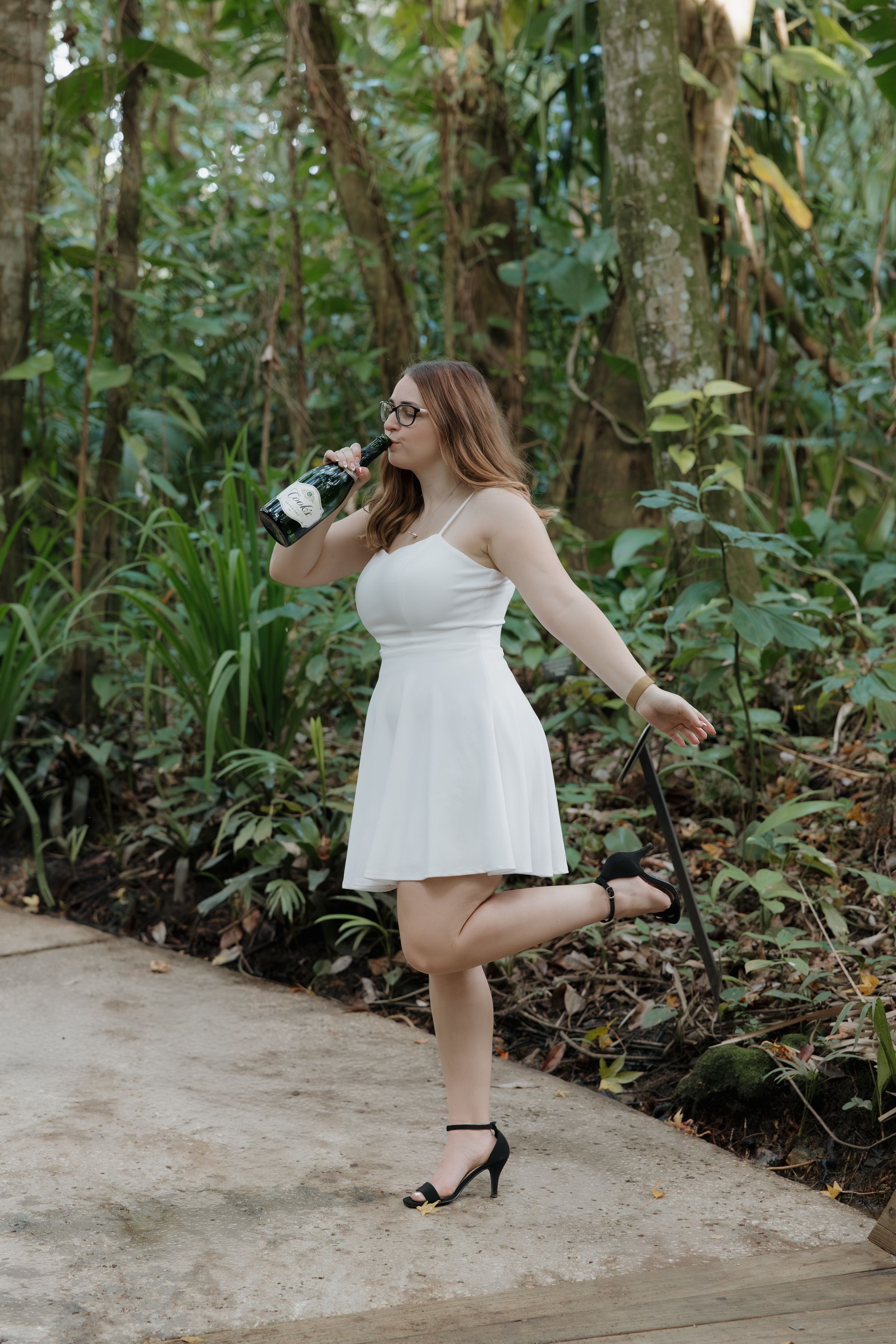Graduate in white dress poses with champagne bottle on tropical garden pathway.