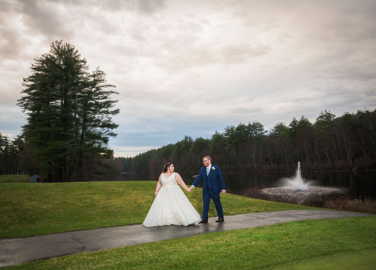 A couple walks along a scenic mountain path surrounded by evergreen trees and cloudy skies.