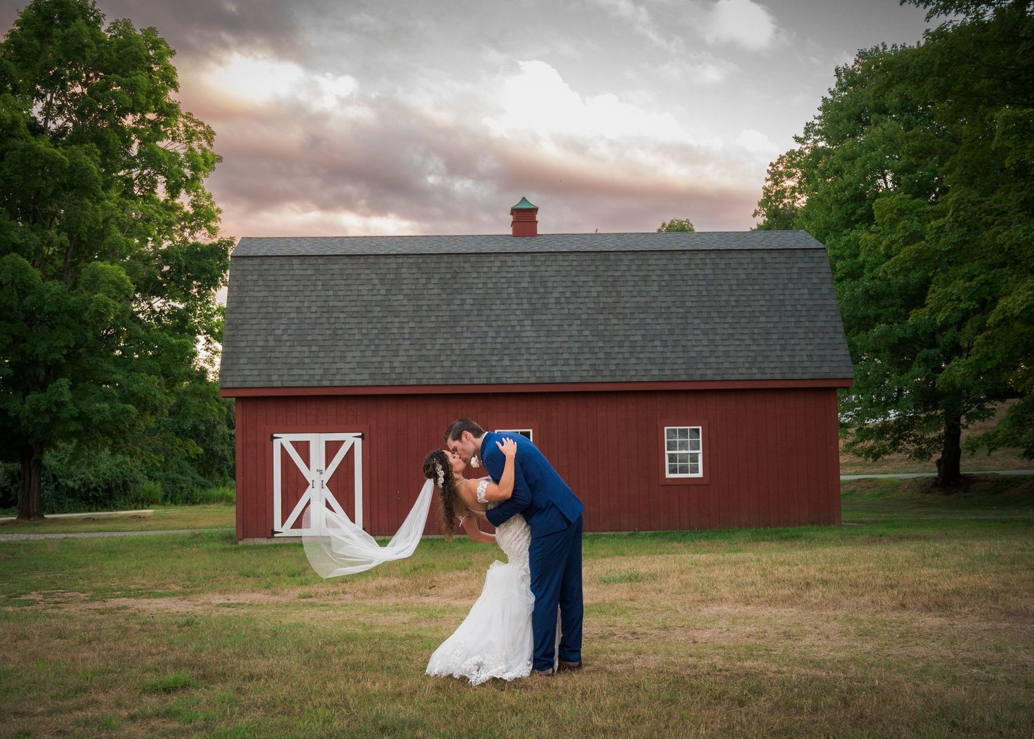 A couple embraces in front of a red barn during a romantic sunset wedding photoshoot.