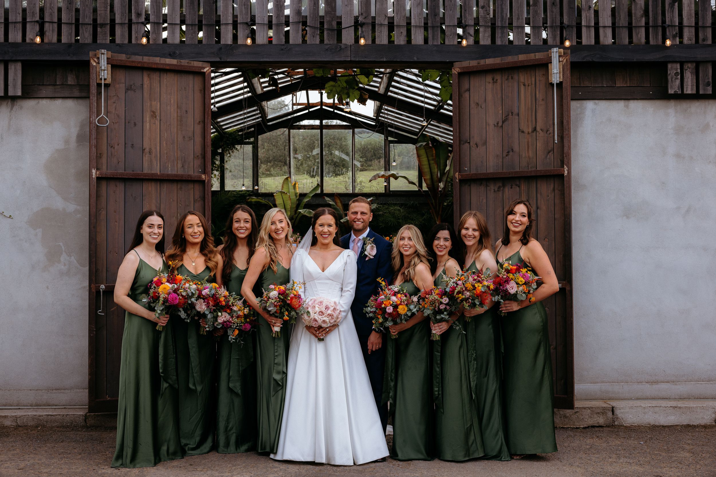 Wedding party lined up in front of rustic barn wearing dark green and white attire.