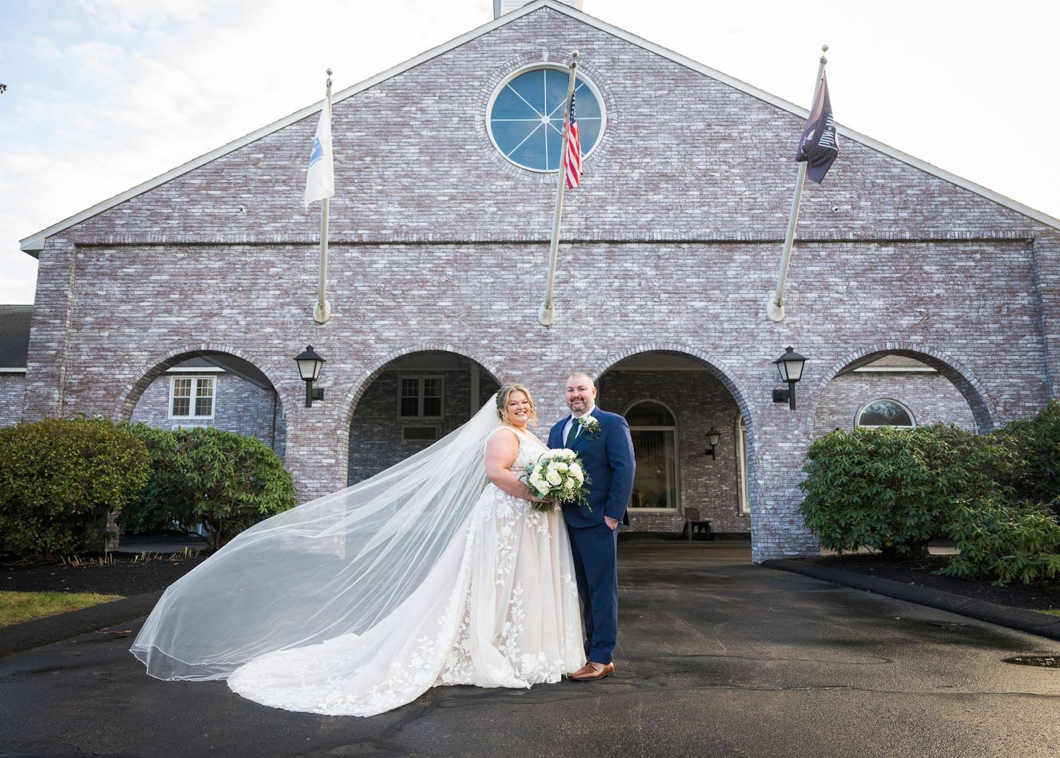 A couple stands outside a stone church building with arched entryways and a flowing white veil catching the breeze.