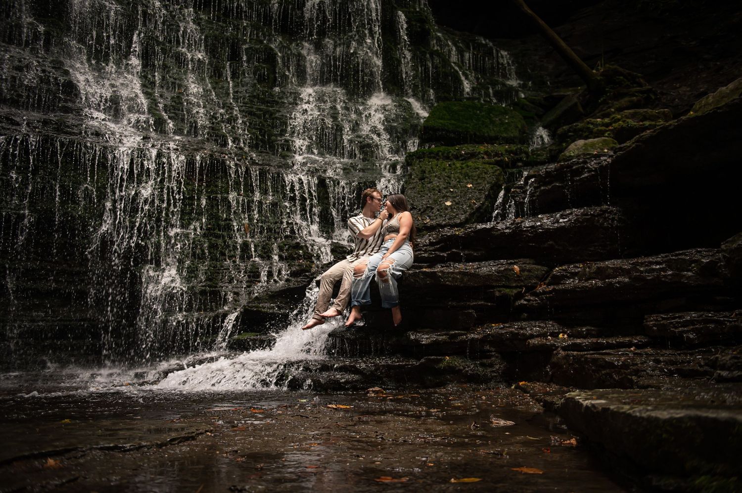 A couple sits in the mist of a dramatic waterfall on wet rocks.