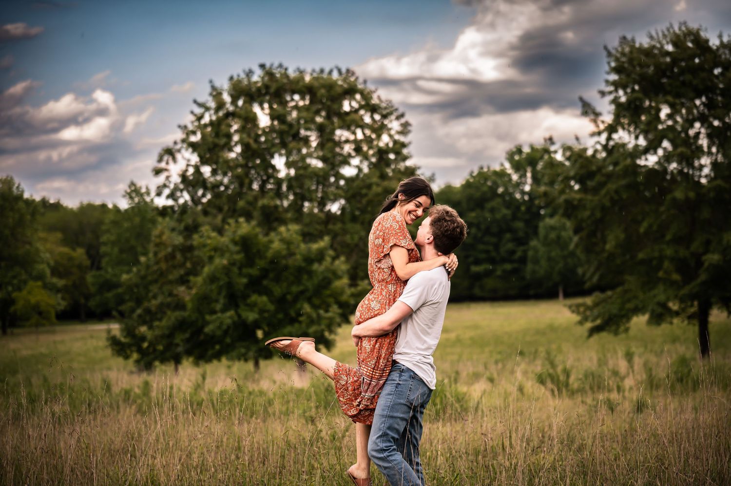 A romantic couple embraces in a sunlit meadow with trees in the background.