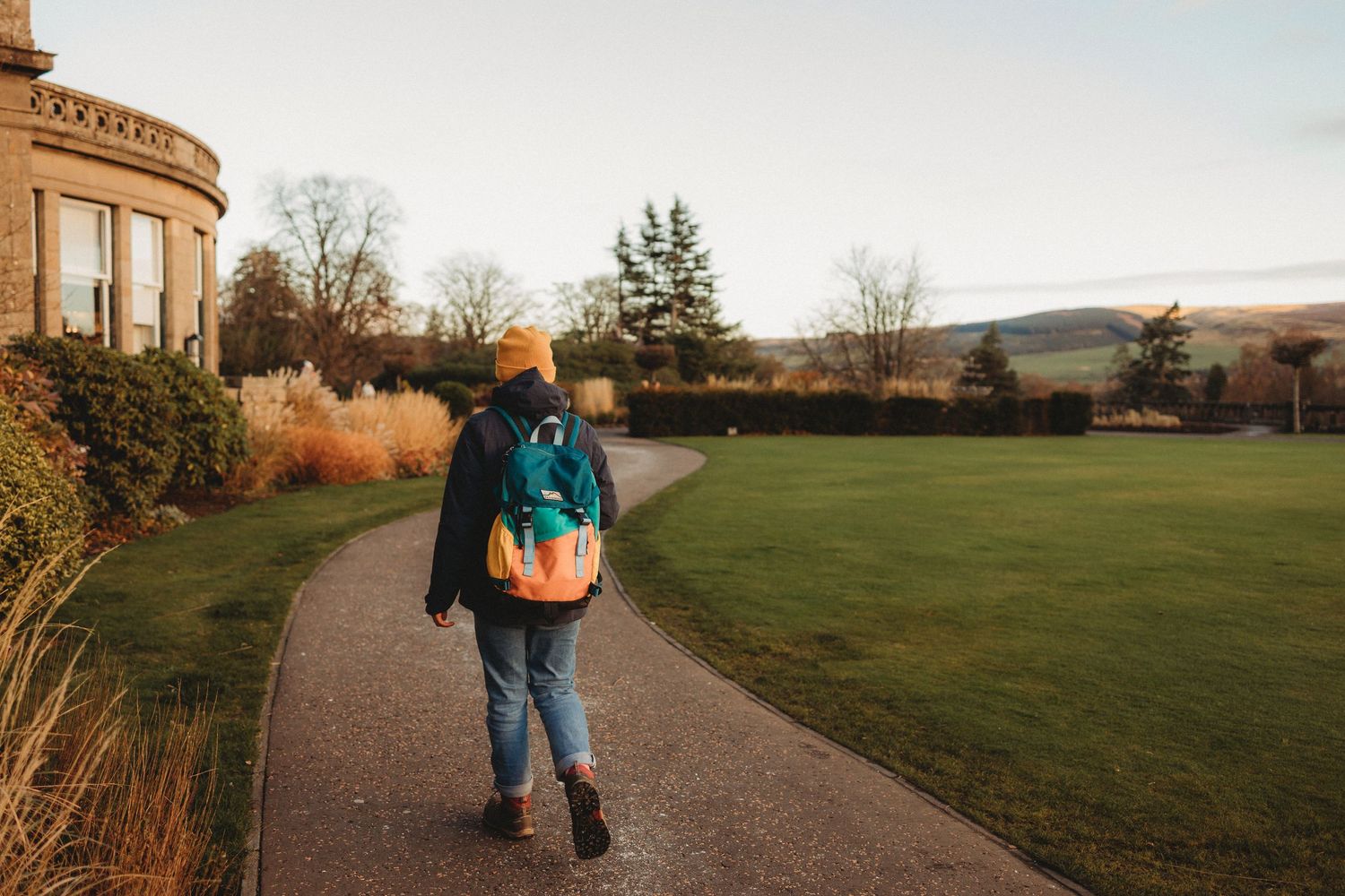 A person with a teal backpack walks along a curved path through manicured grounds at sunset.