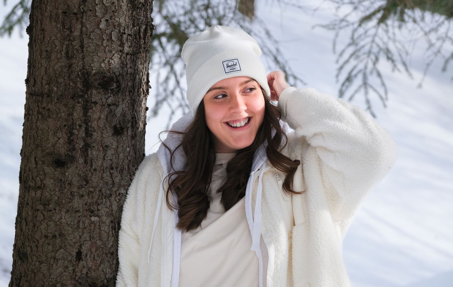Person in white winter coat and beanie hat smiling by tree in snowy setting.
