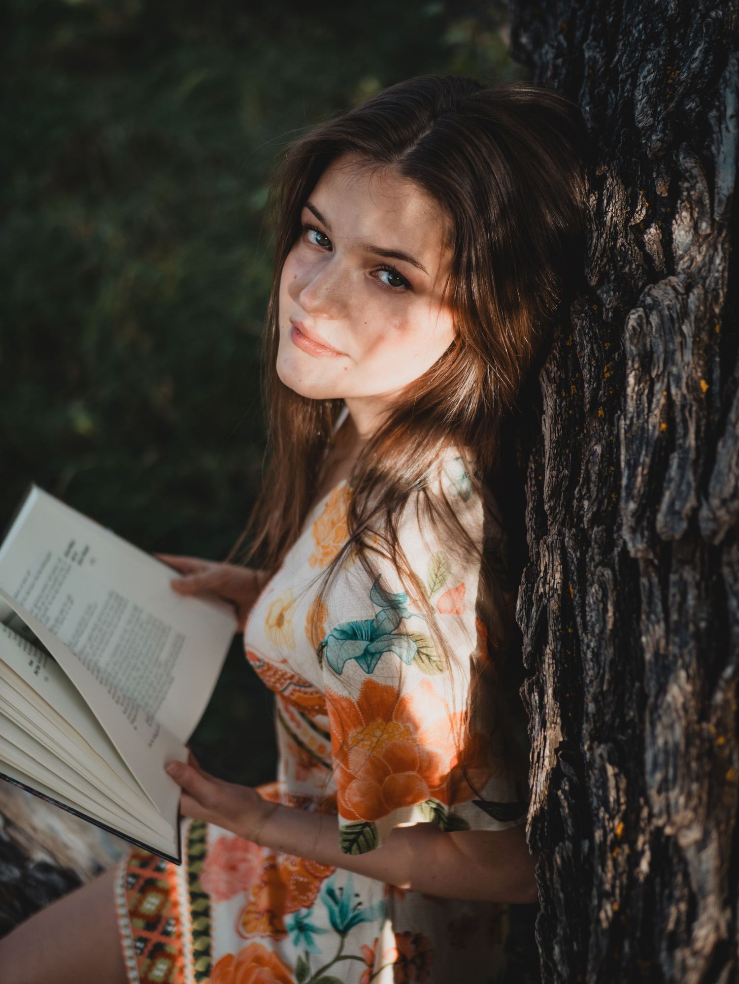 A person in a floral dress reads a book against a dark tree trunk in dappled sunlight.
