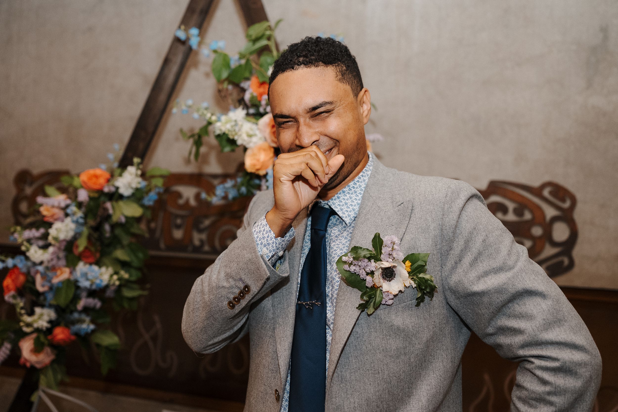 A groom in a gray suit and blue tie laughs while getting ready for his wedding celebration.