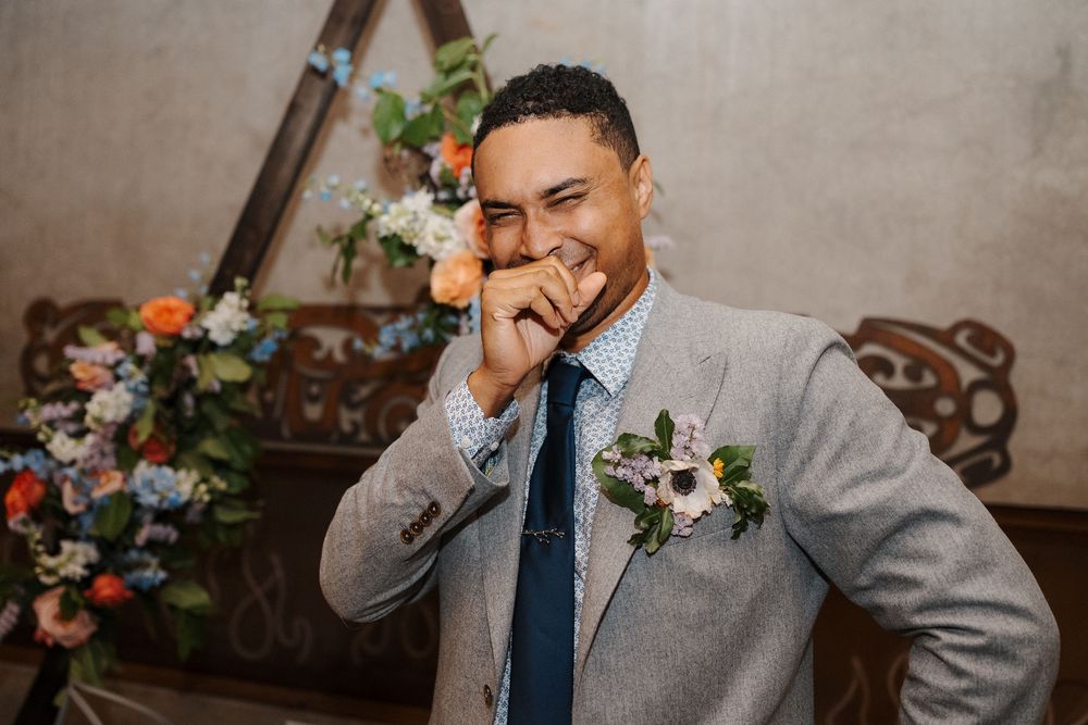 A groom in a gray suit and blue tie laughs while getting ready for his wedding celebration.