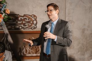 Professional in a gray suit and blue tie holds a cup while standing against a textured wall.